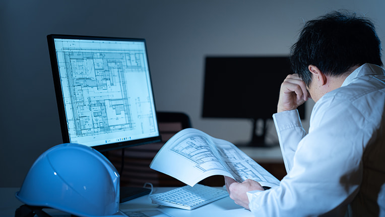 深夜にパソコンと紙で図面を見て悩む作業着姿の施工管理男性