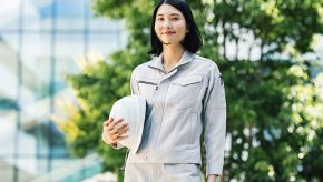 ビルの前でヘルメットを持って笑顔で立つ作業着姿の女性施工管理